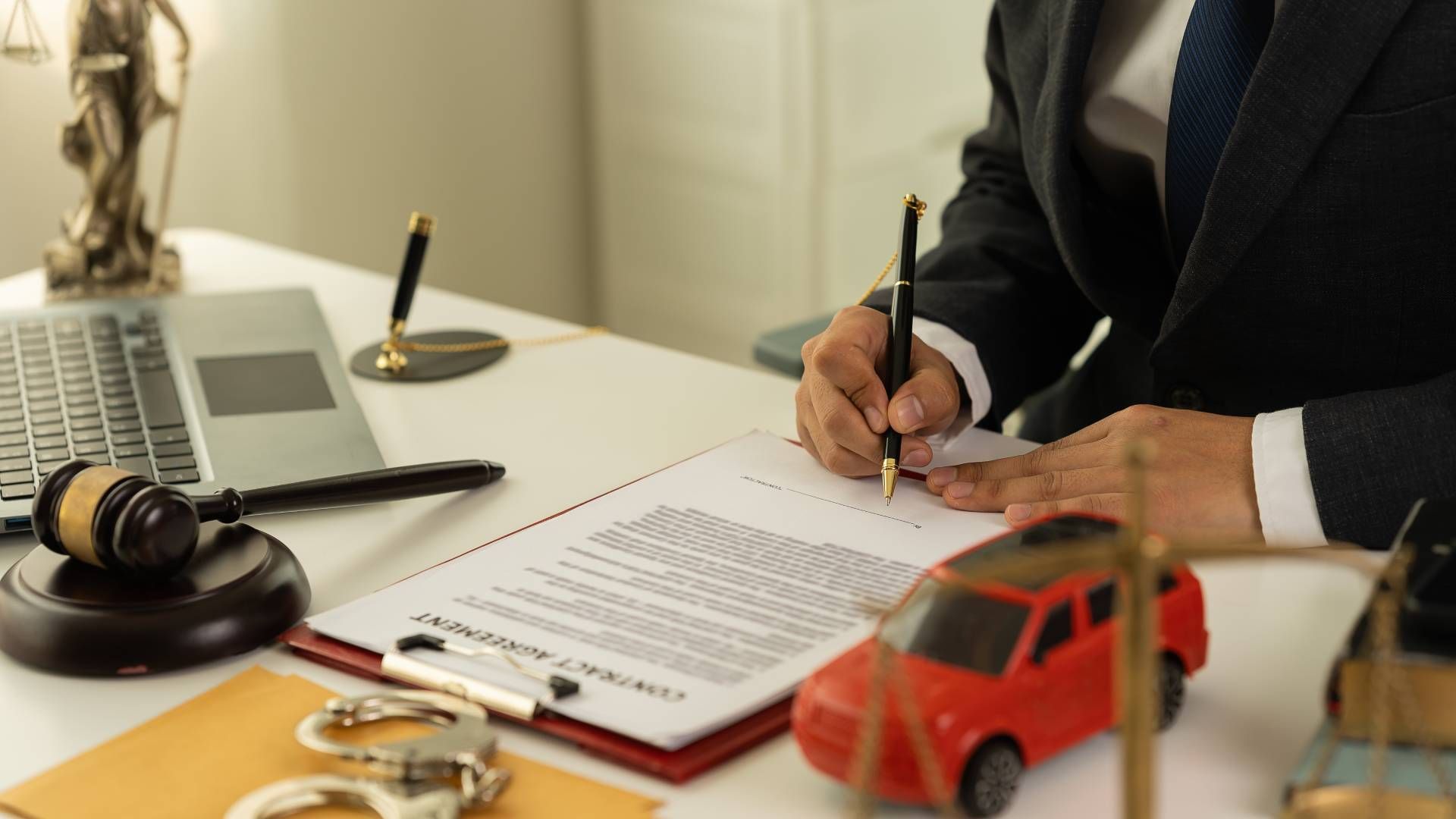 Person signing contract at desk with toy car, laptop, gavel, handcuffs, and statue of justice.