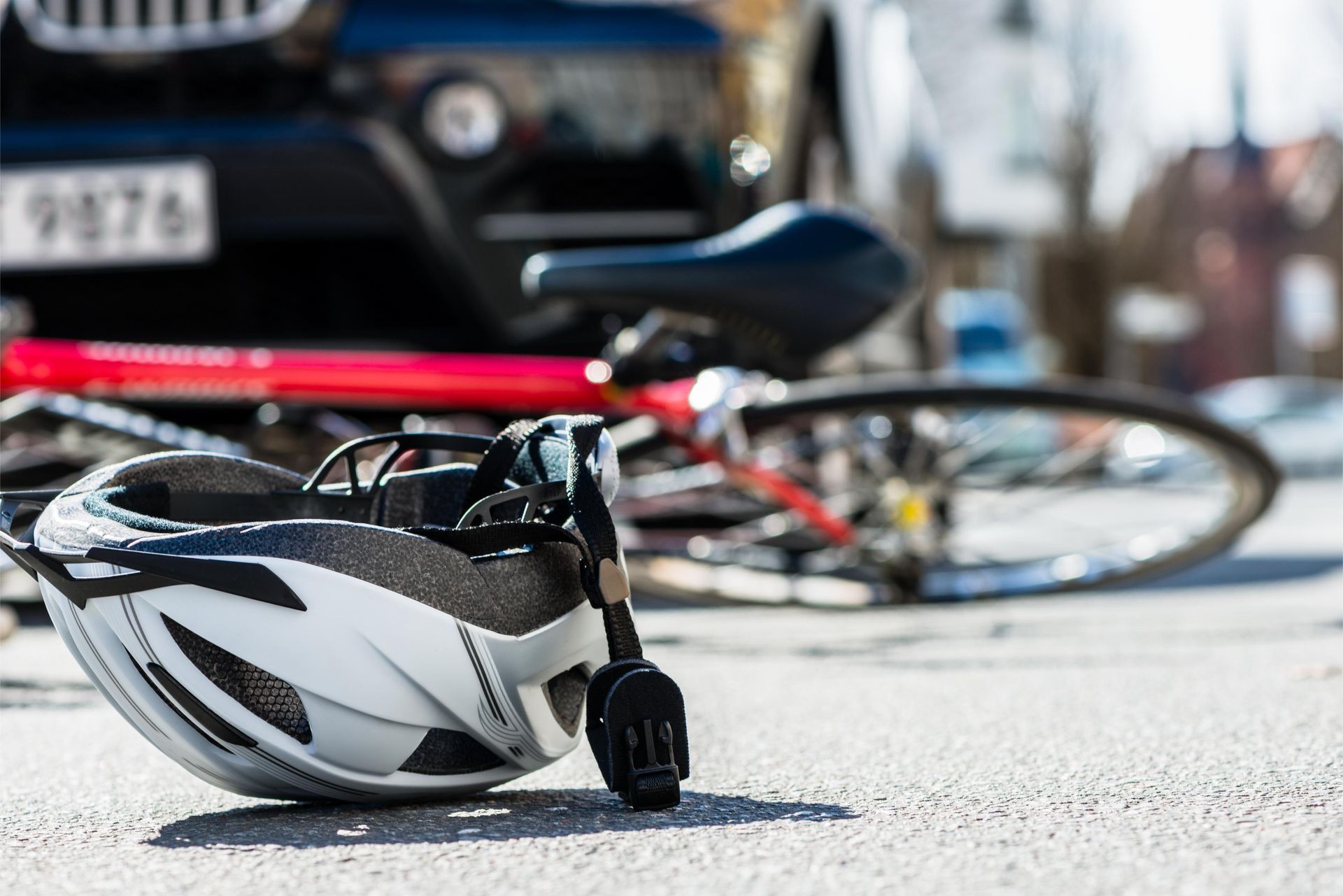 A white bicycle helmet rests on the asphalt near a red bicycle lying on the ground in front of a car.