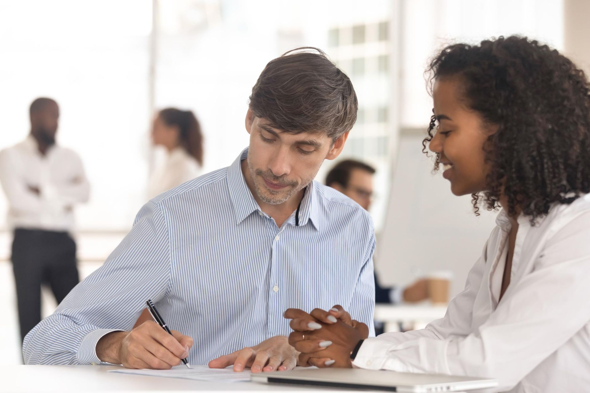 Two colleagues discuss and write on a document at an office desk, with others working in the blurred background.