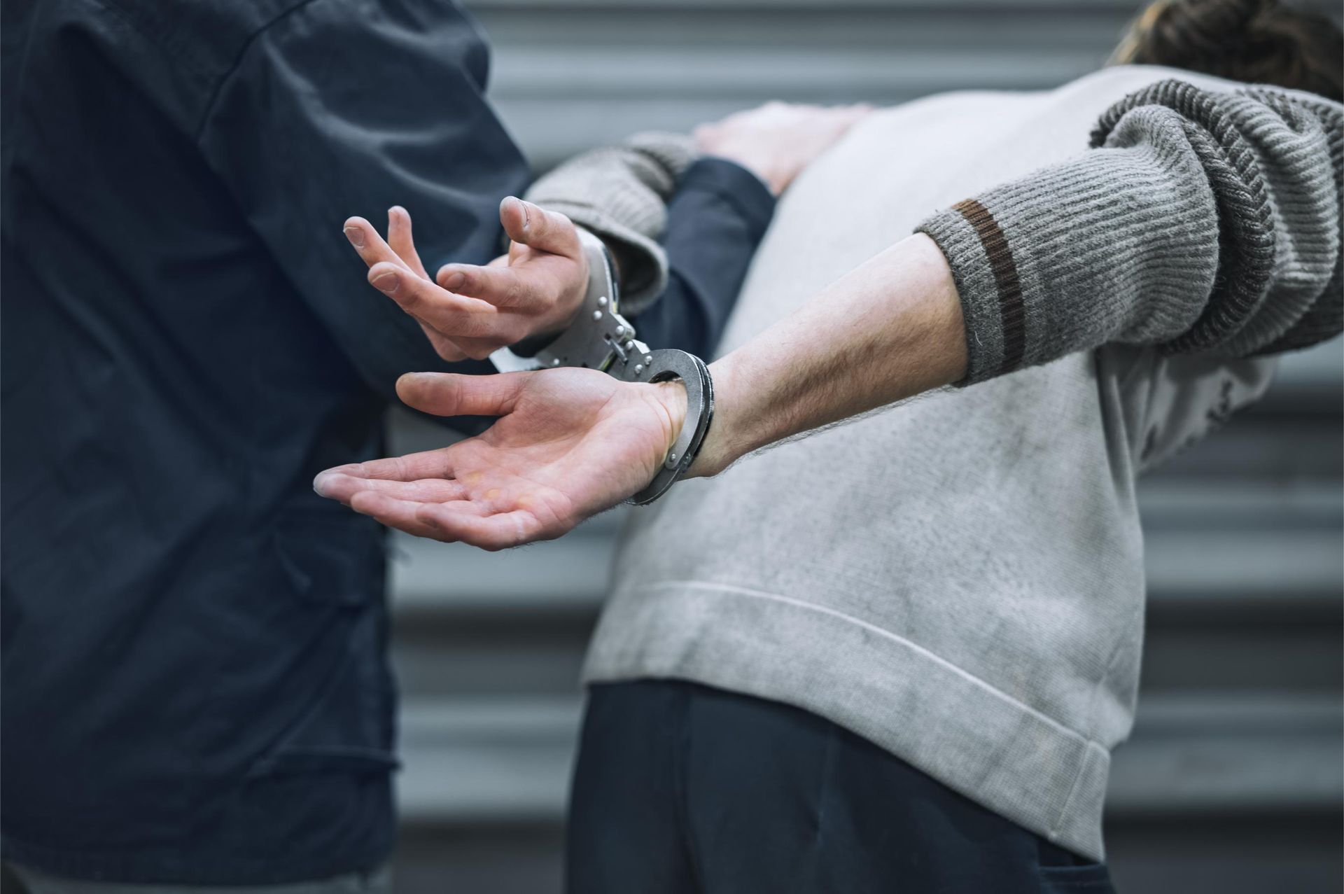 A person in a gray sweater stands with their hands behind their back, held in handcuffs by another person in a dark jacket.
