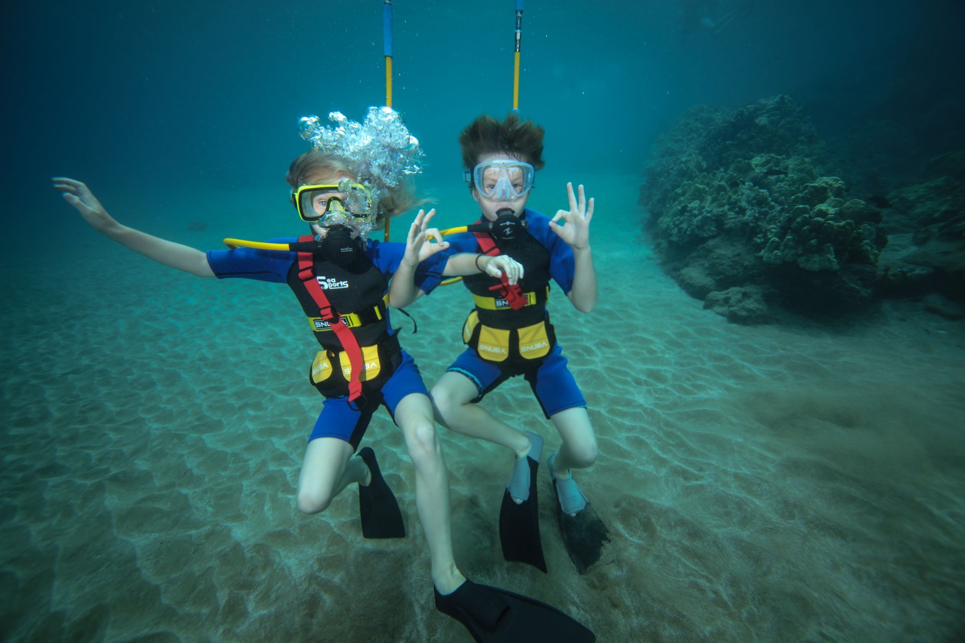 Two children are scuba diving in the ocean and giving the ok sign.