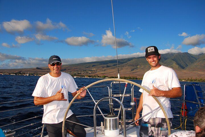 Two men are sitting at the helm of a sailboat giving a thumbs up