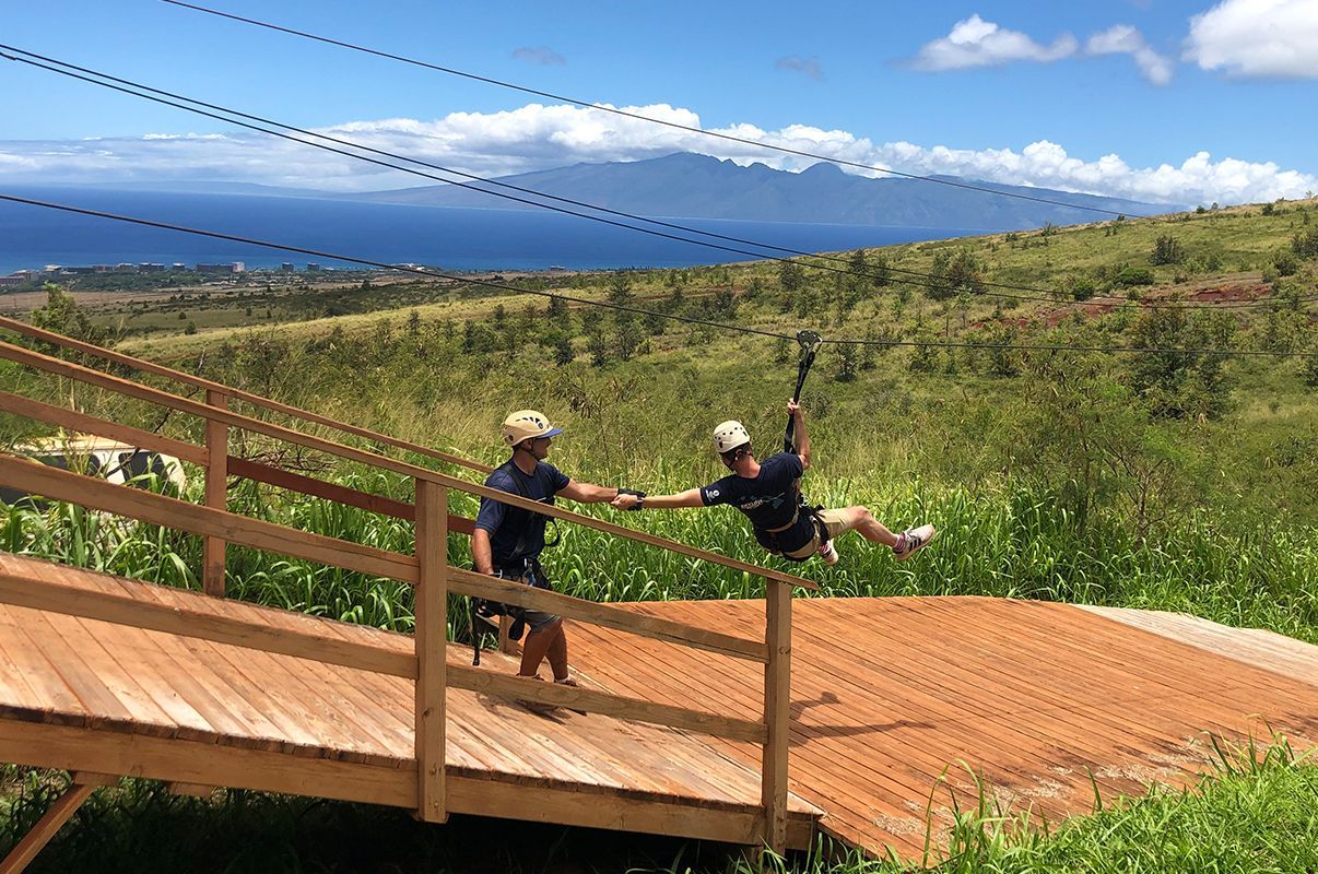 Two people are riding a zip line over a wooden bridge.