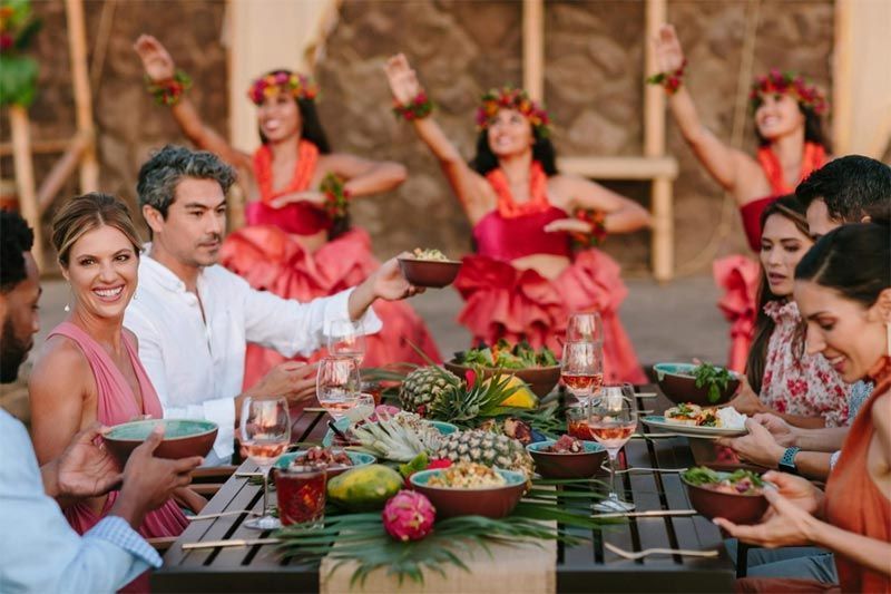 A group of people are sitting at a table with bowls of food and hula dancers in the background.