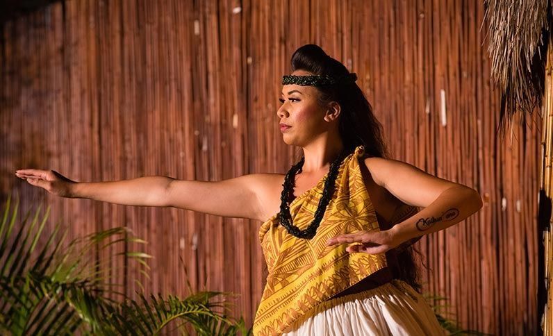 A woman is dancing a hula in front of a wooden wall.