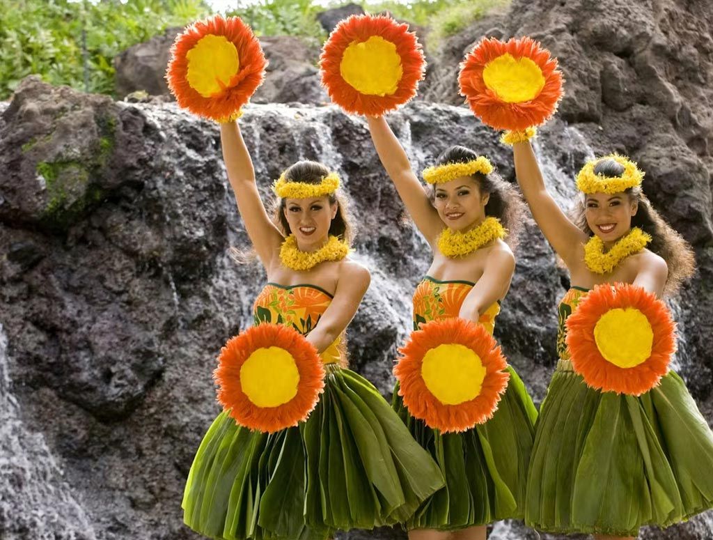 Three hula dancers are holding up flowers in front of a waterfall