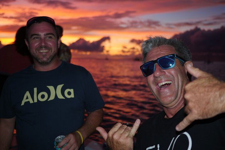 Two men wearing aloha shirts are posing for a picture in front of a sunset.