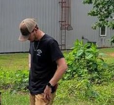 A man wearing a hat is standing in the grass in front of a building.