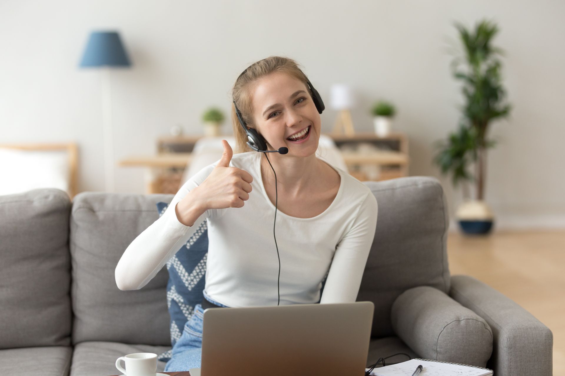 A woman is sitting on a couch with a laptop and giving a thumbs up.