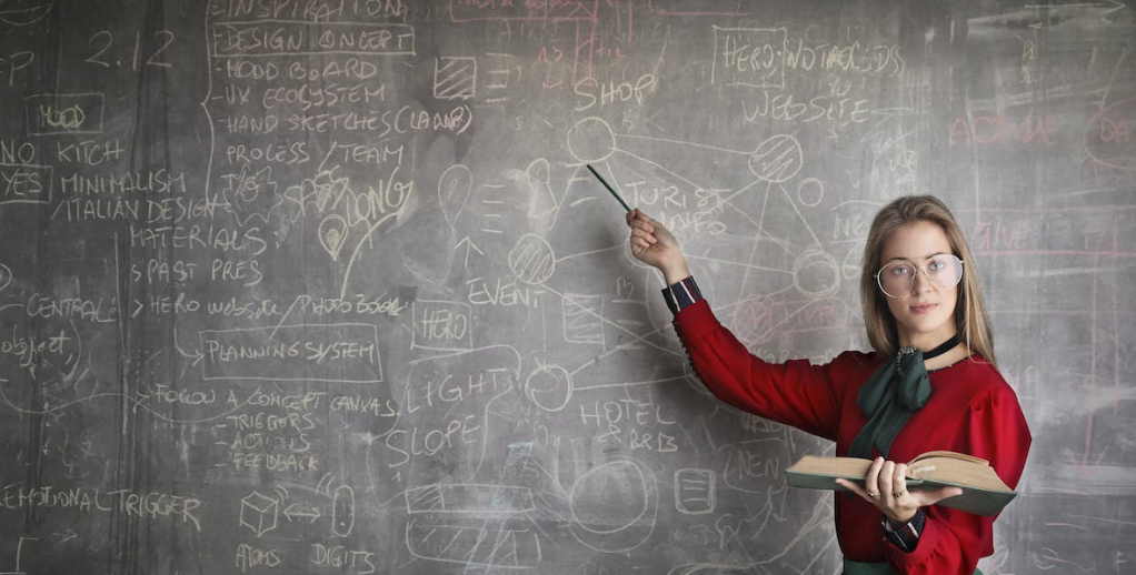 A woman is standing in front of a blackboard holding a book and pointing at it.