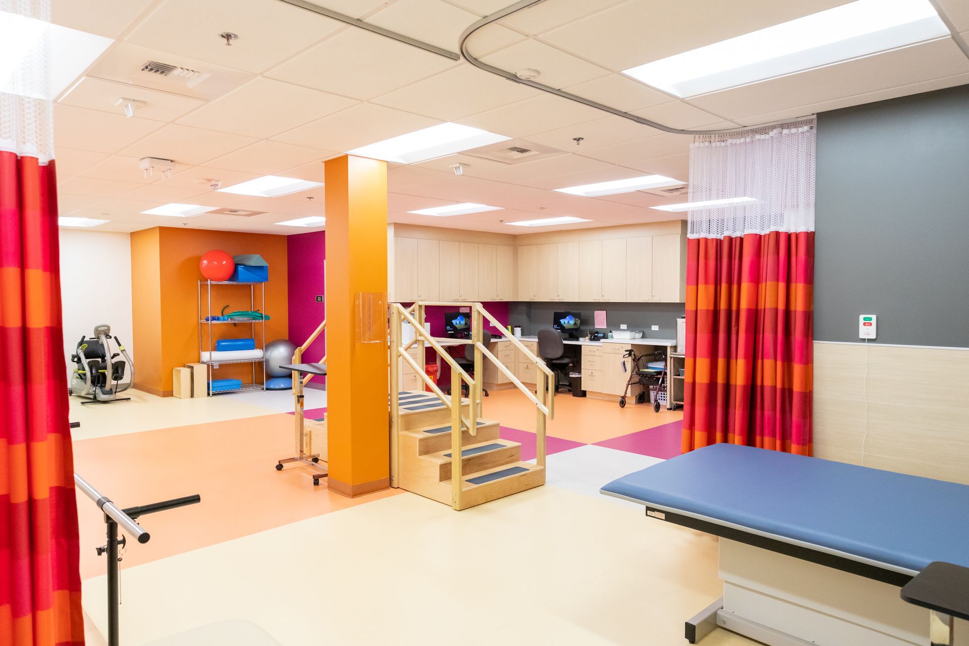 Physical therapy room with orange, pink, and blue accents. Features stairs, equipment, and a treatment table.