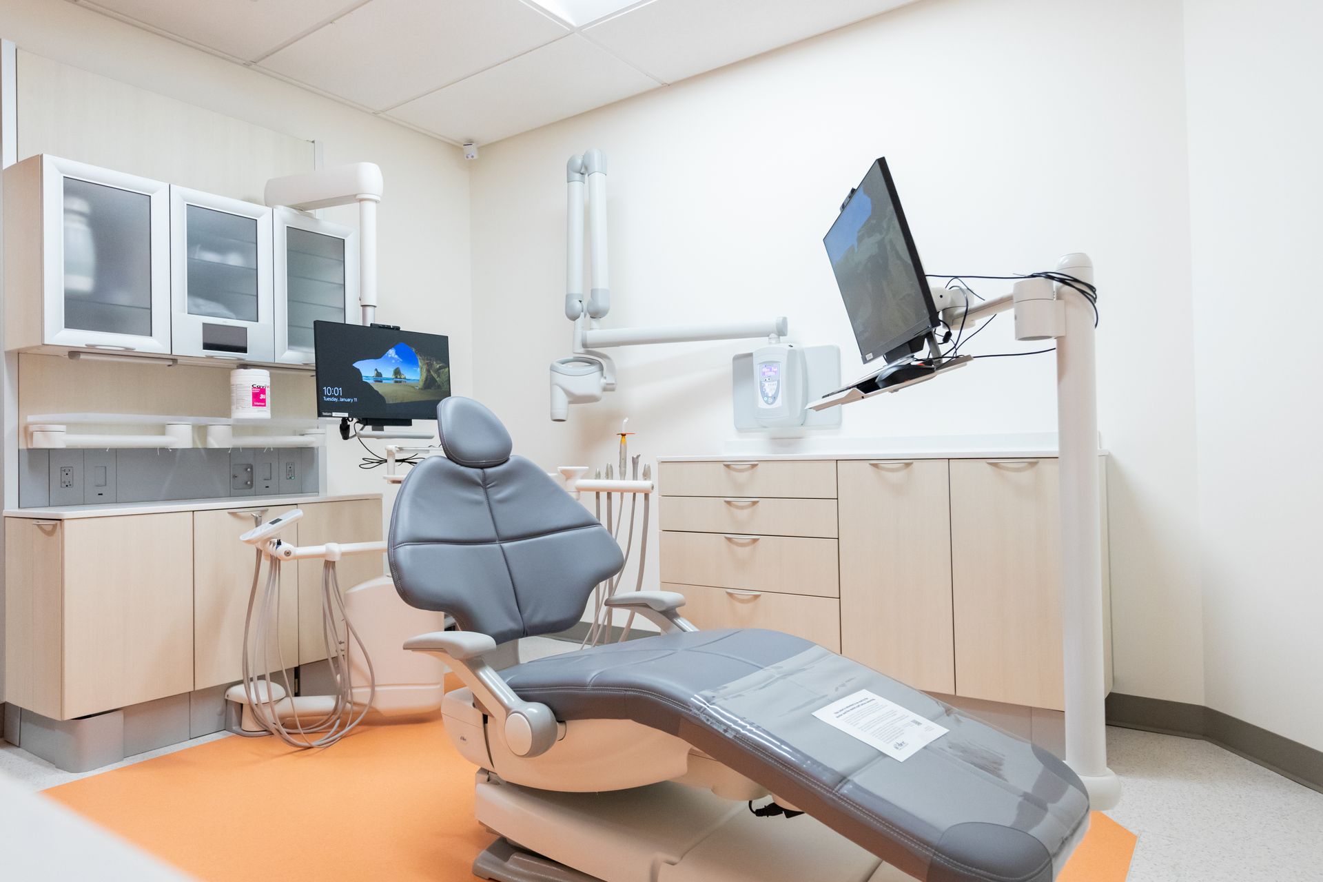 A dental office exam room with a gray chair, monitors, and beige cabinets.
