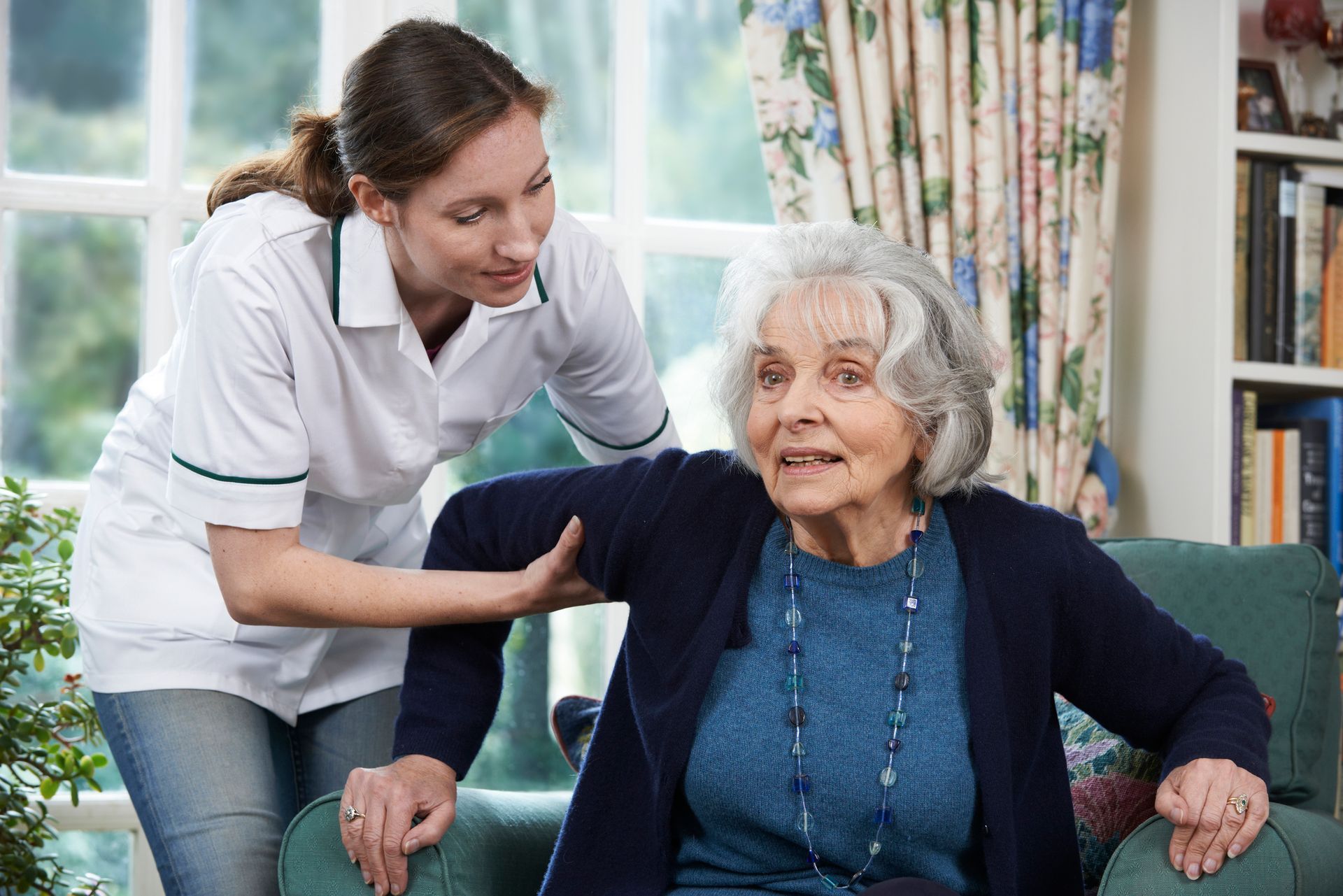 Female caregiver in uniform assisting elderly woman seated in armchair at home.
