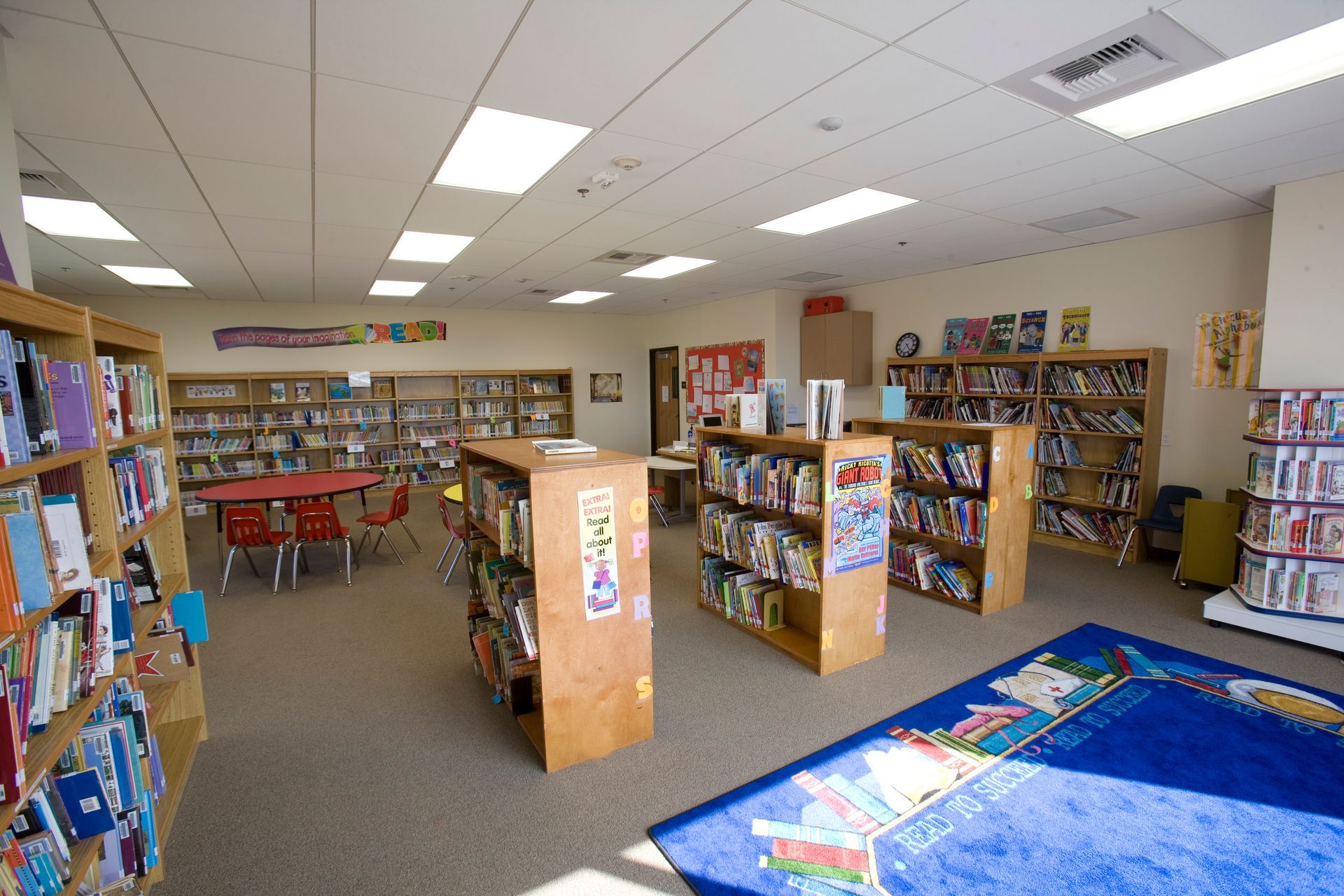 Interior of a library with bookshelves, a reading rug, and a round table with chairs for children.