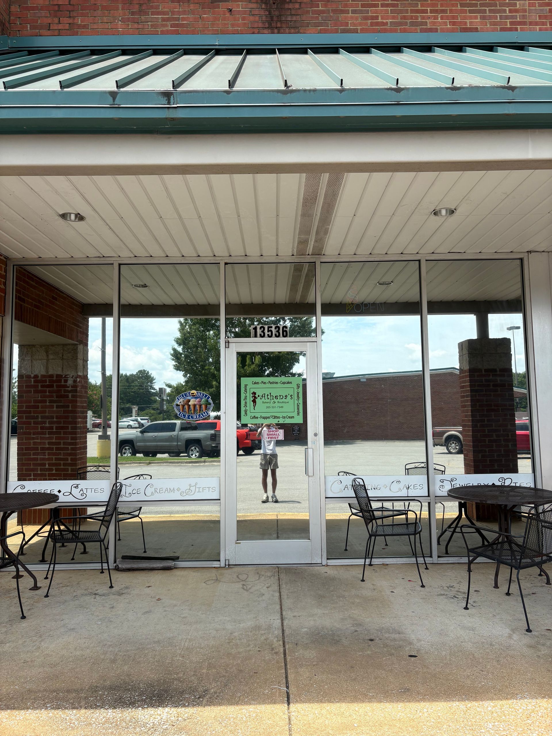 Exterior of a business with glass front. A person is visible inside. Tables and chairs are outside.