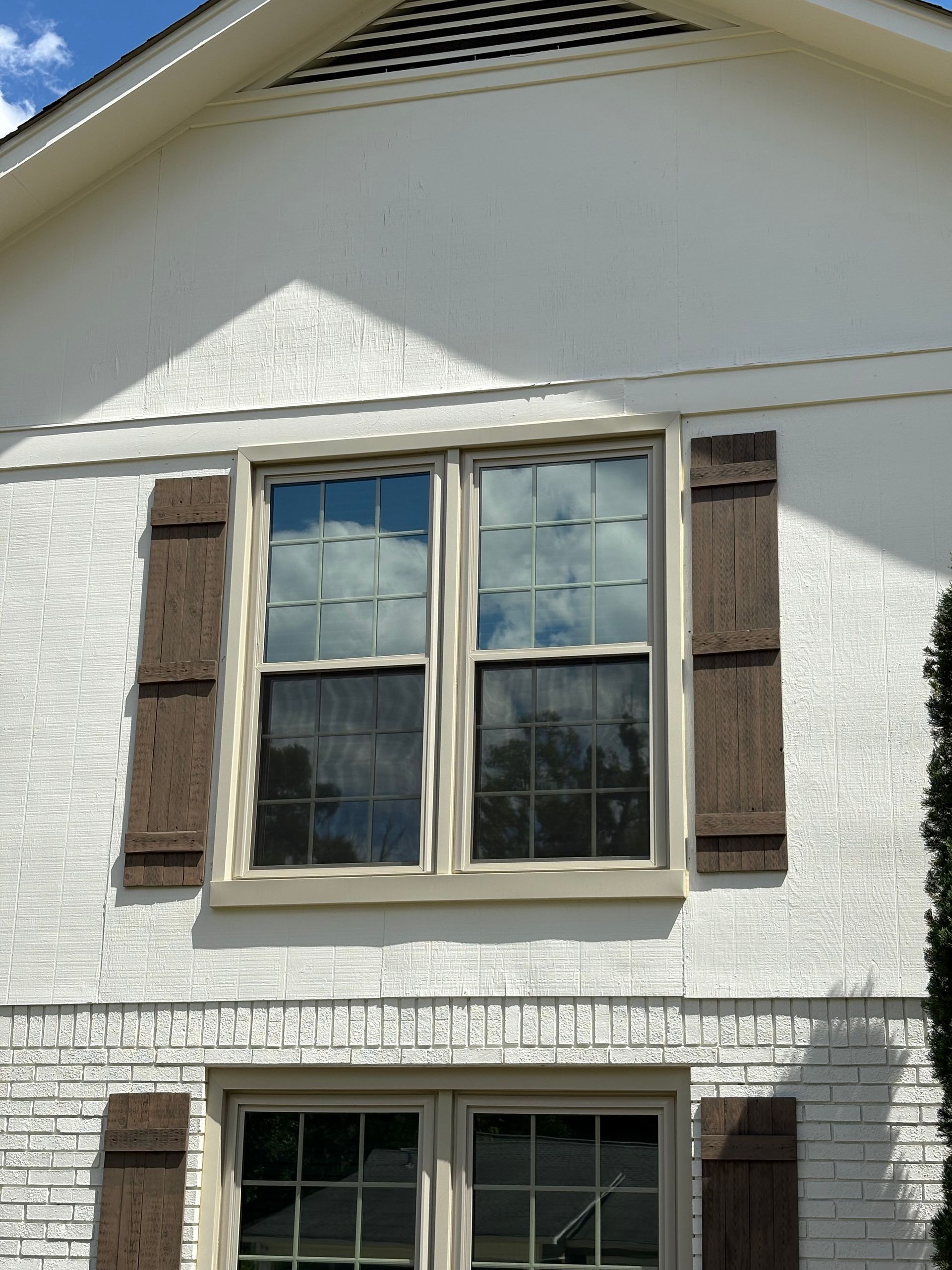 Two story house with brown shutters on white brick and stucco. Window reflections of sky.