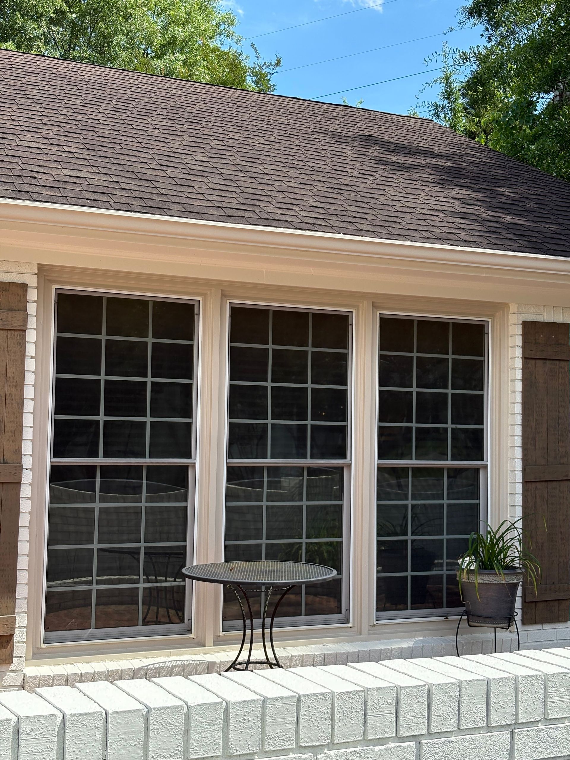 Three-pane window with dark screens, set in a light beige frame, above a brick ledge. Table and potted plant on ledge.