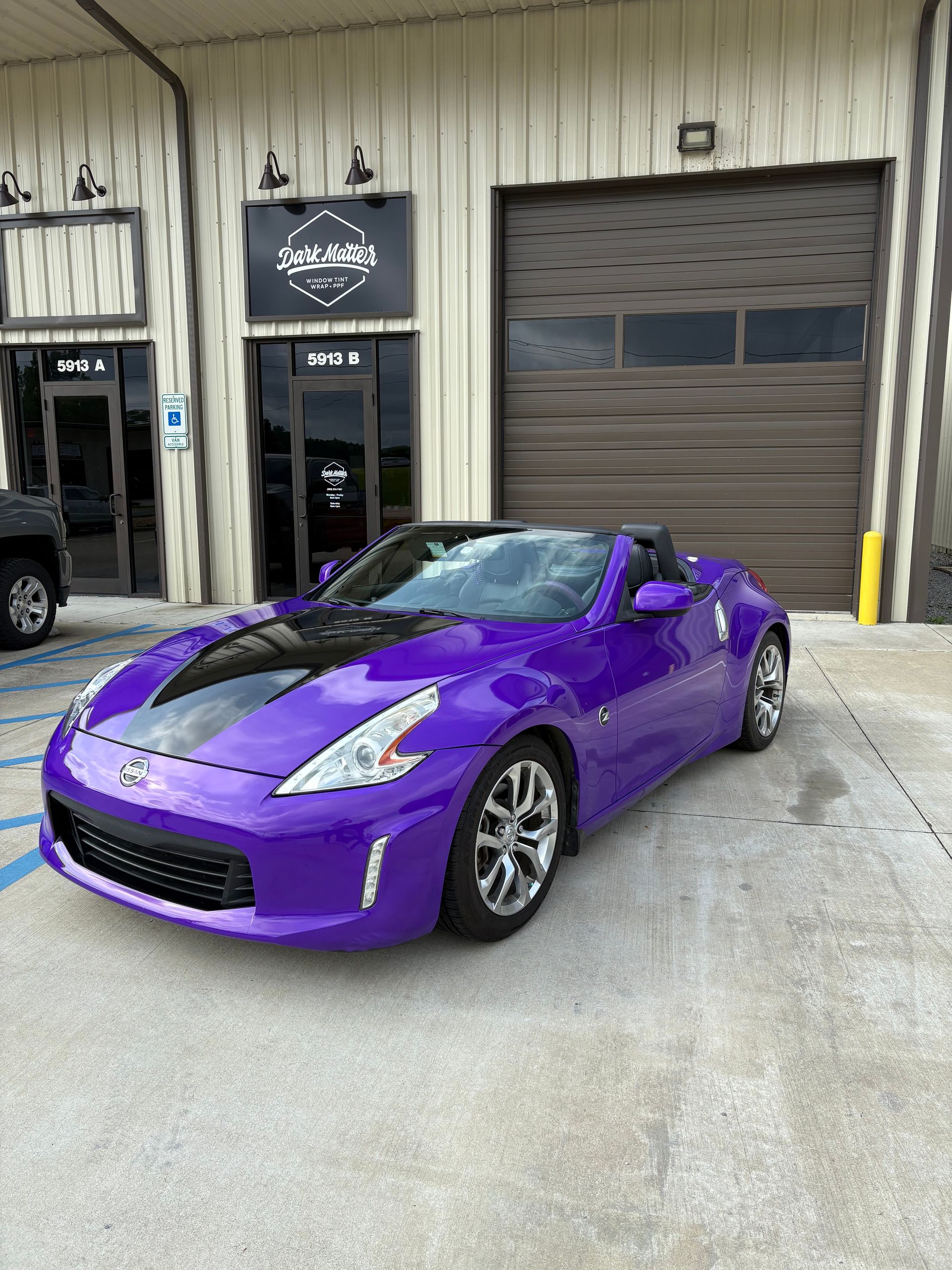 Purple Nissan 370Z convertible with a black hood parked outside a building with a garage door.