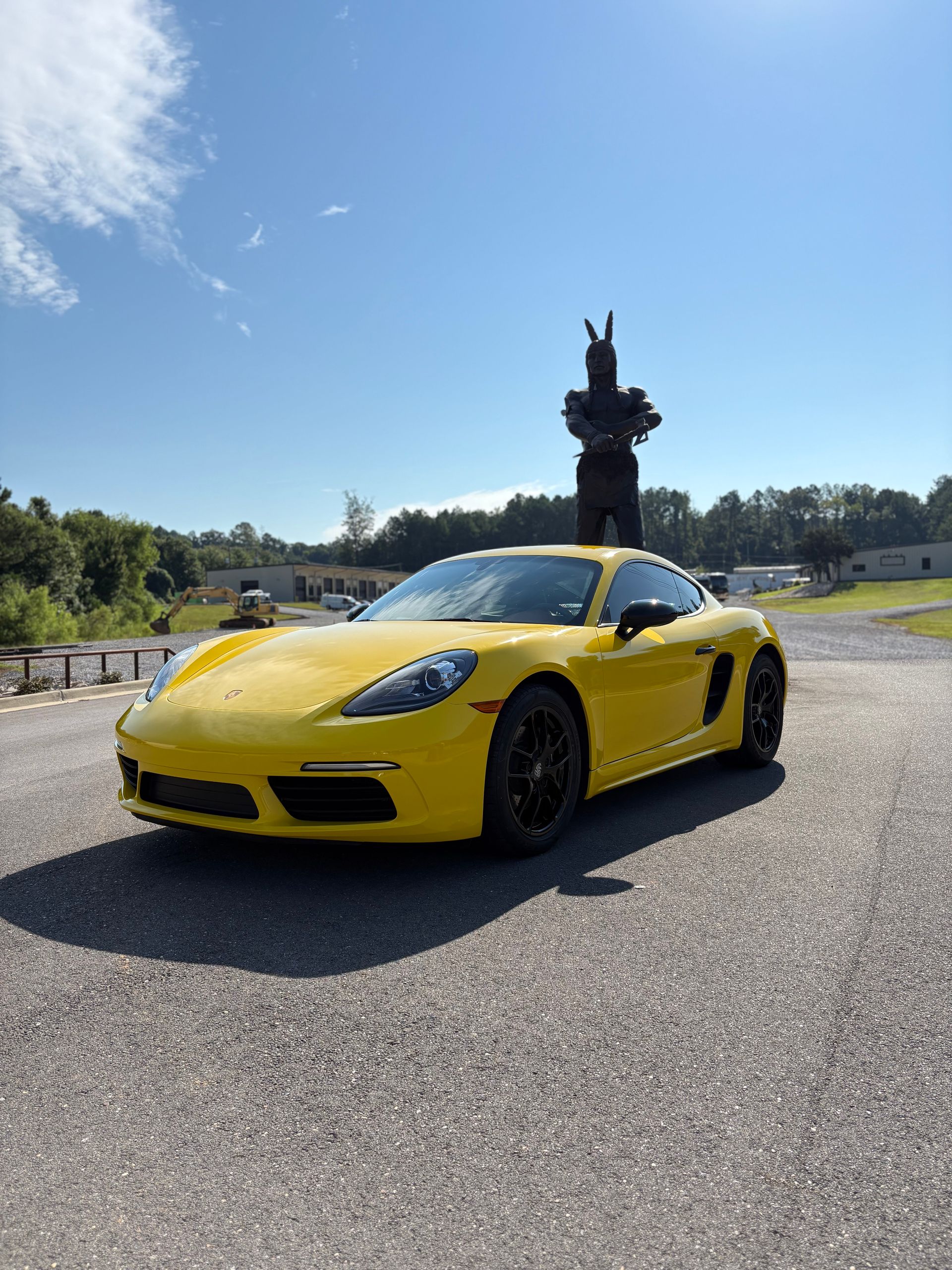 Yellow Porsche sports car parked on a paved lot on a sunny day with a monument in the background.