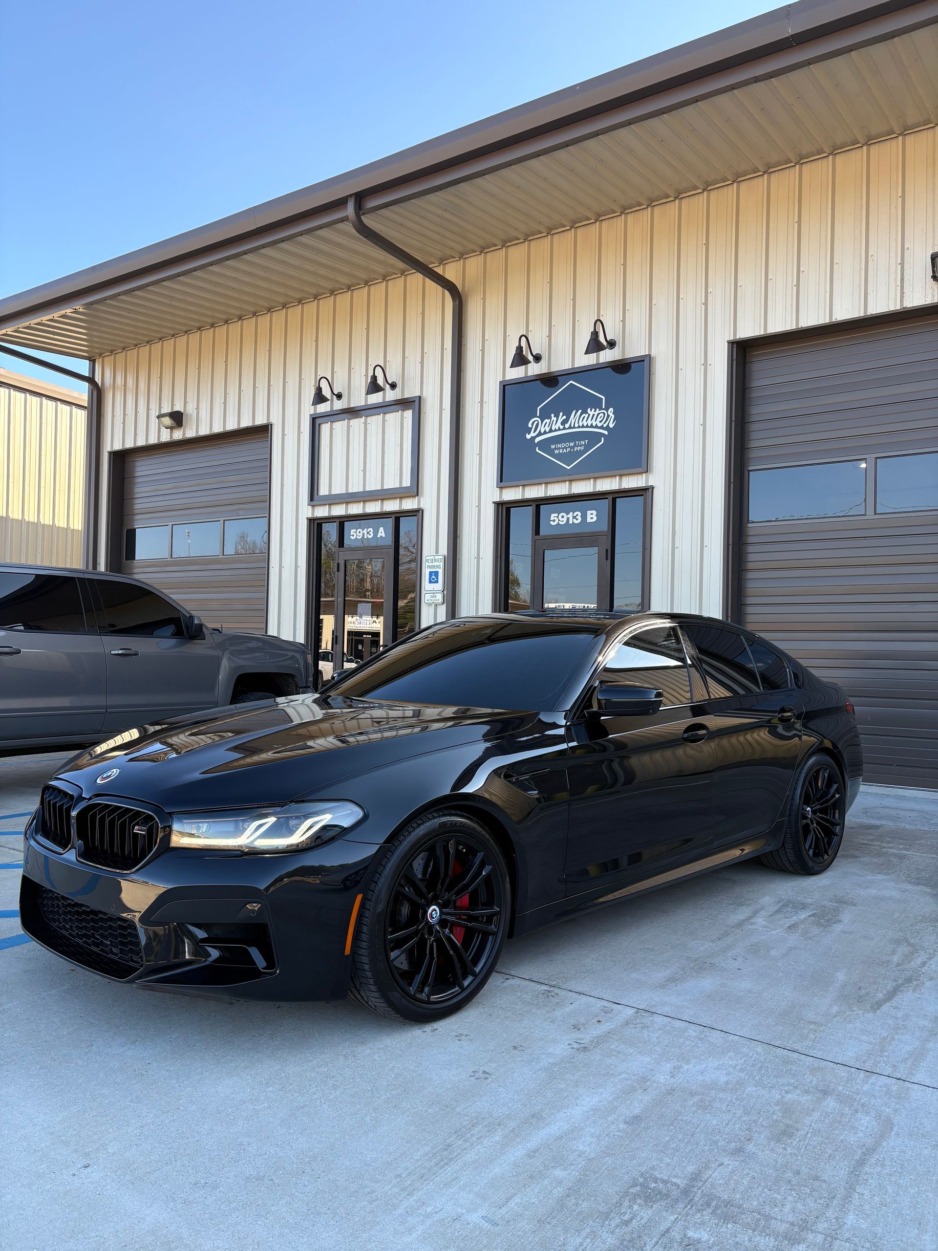 Black BMW M5 parked in front of a building with glass doors and overhead lights.