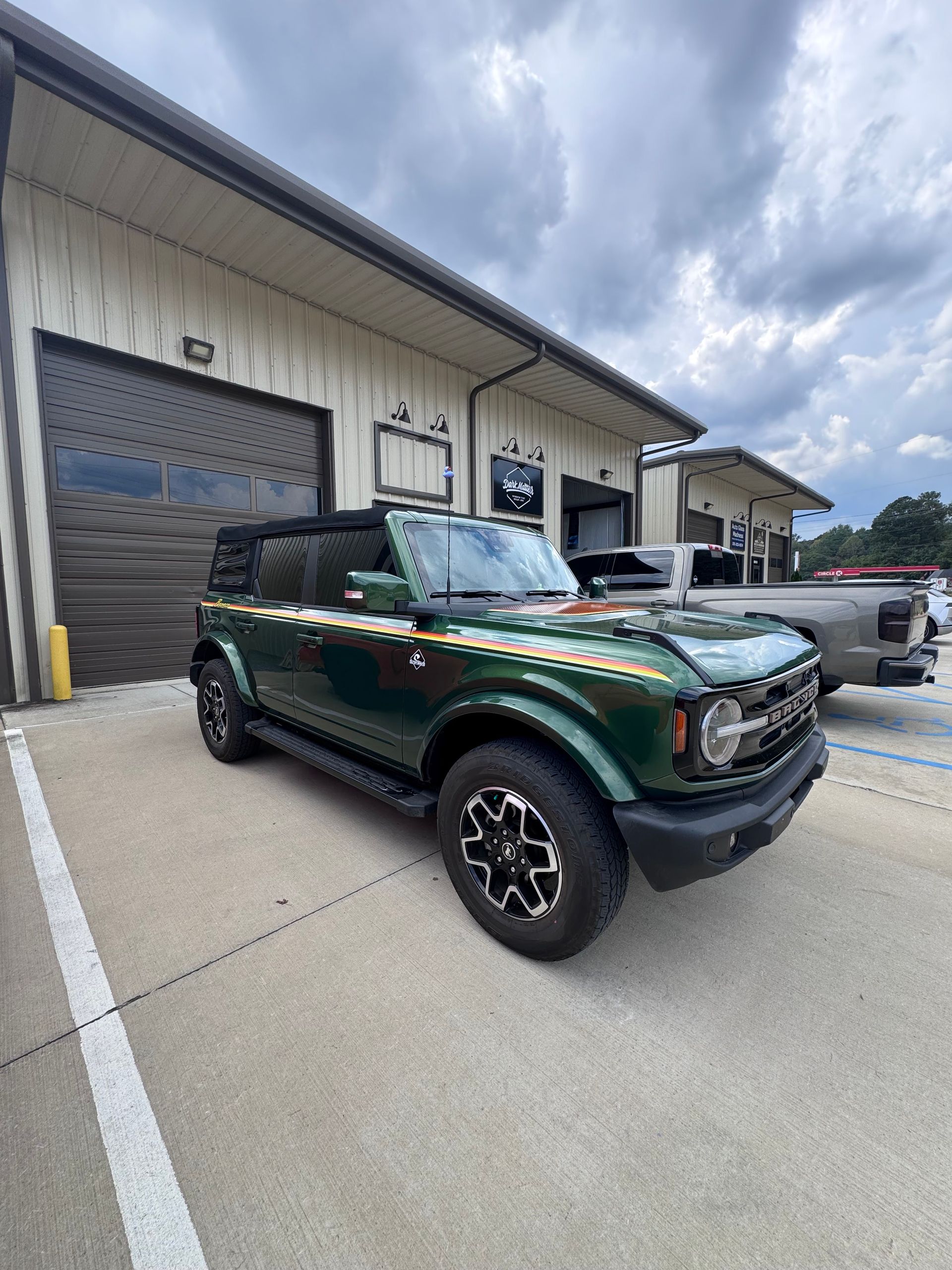Green Ford Bronco parked in front of a building with a roll-up door. Gold and white side stripes.