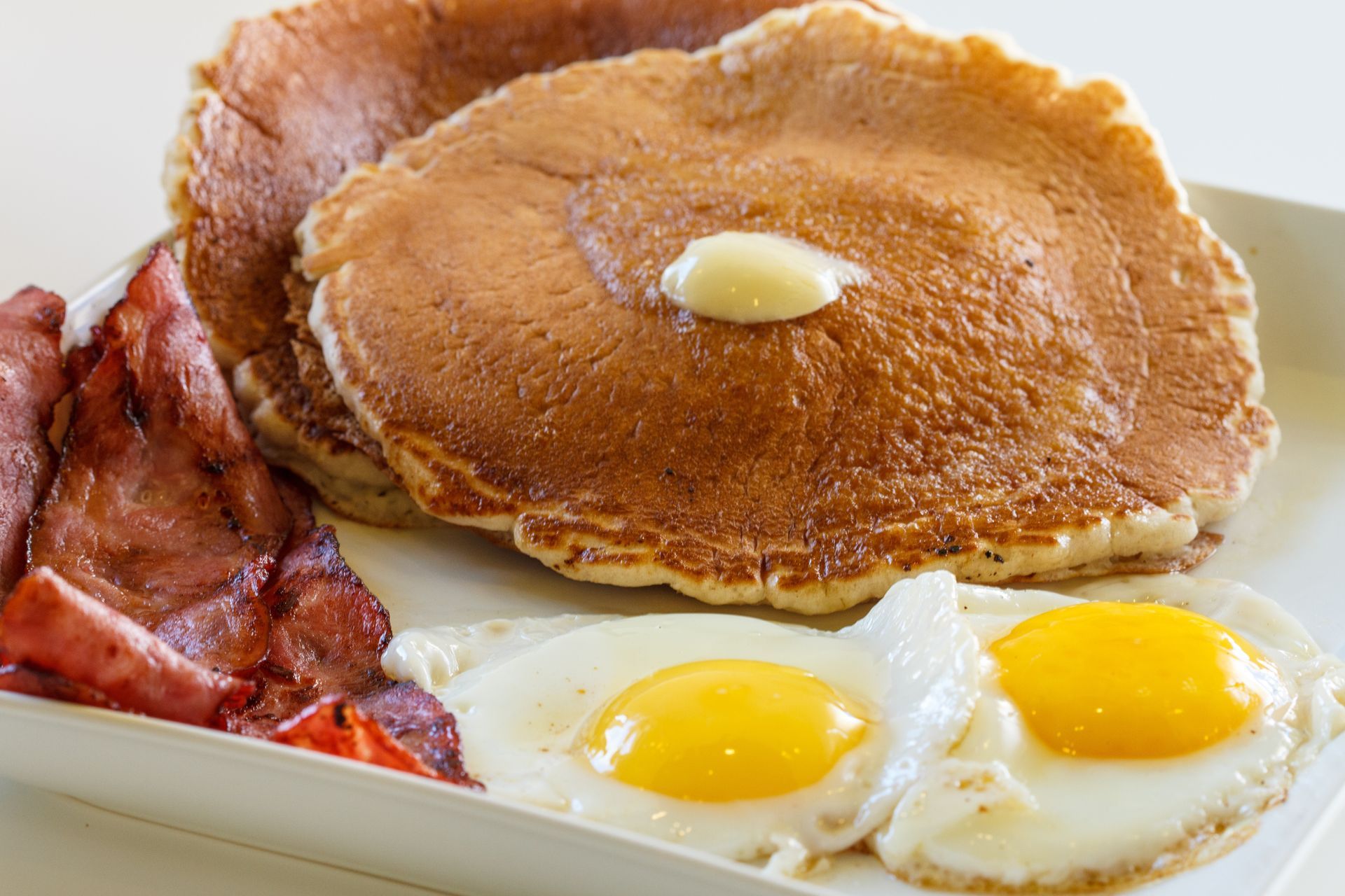 A breakfast plate featuring a stack of pancakes with a pat of butter, several strips of bacon, and two sunny-side up eggs.