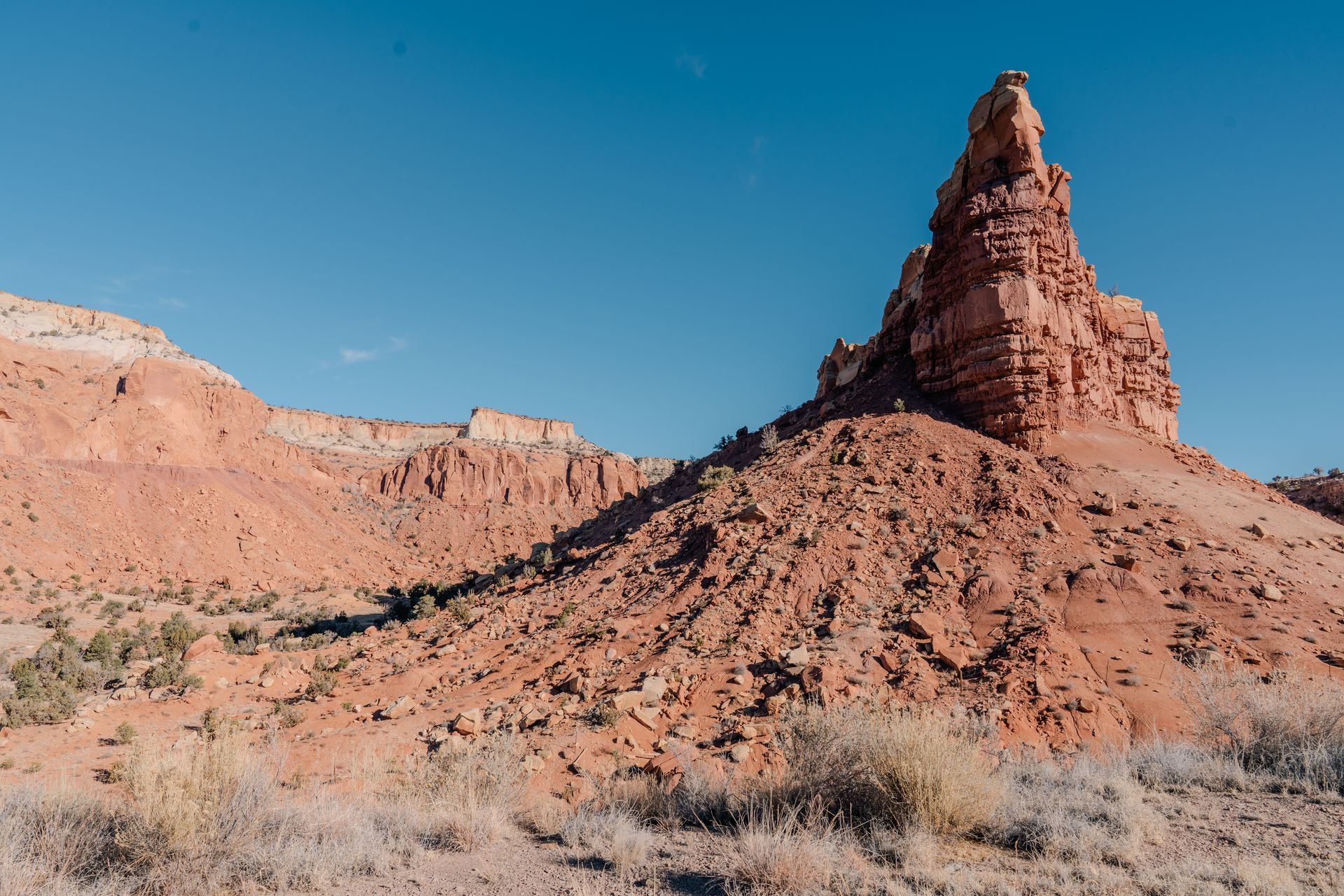 A tall, jagged red rock spire rises above a dusty, sparse desert landscape under a clear blue sky.