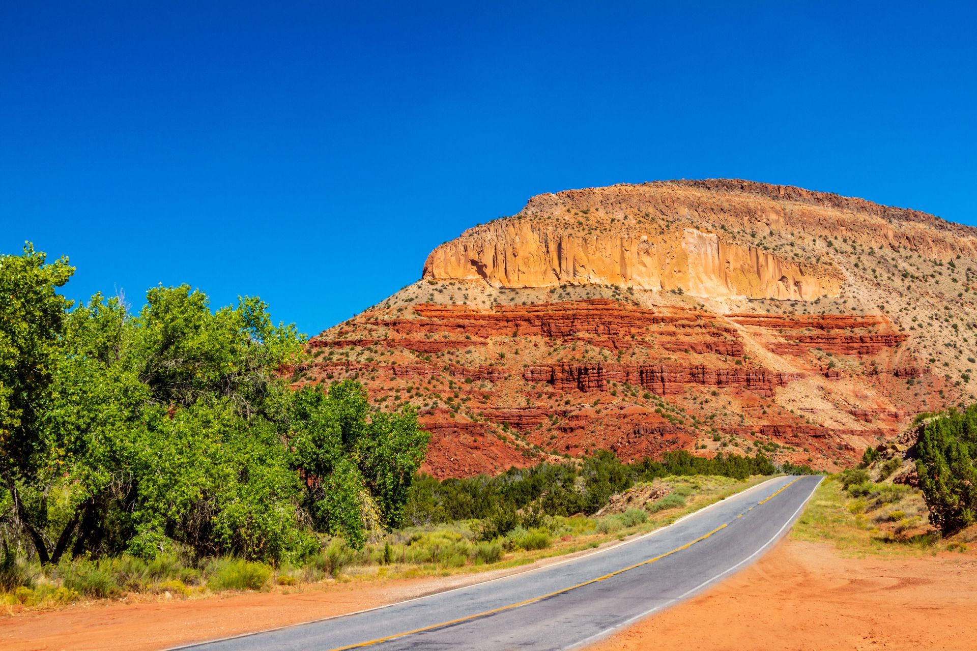 A paved road curves toward a large, layered red rock mesa under a bright blue sky, with green trees along the left side.