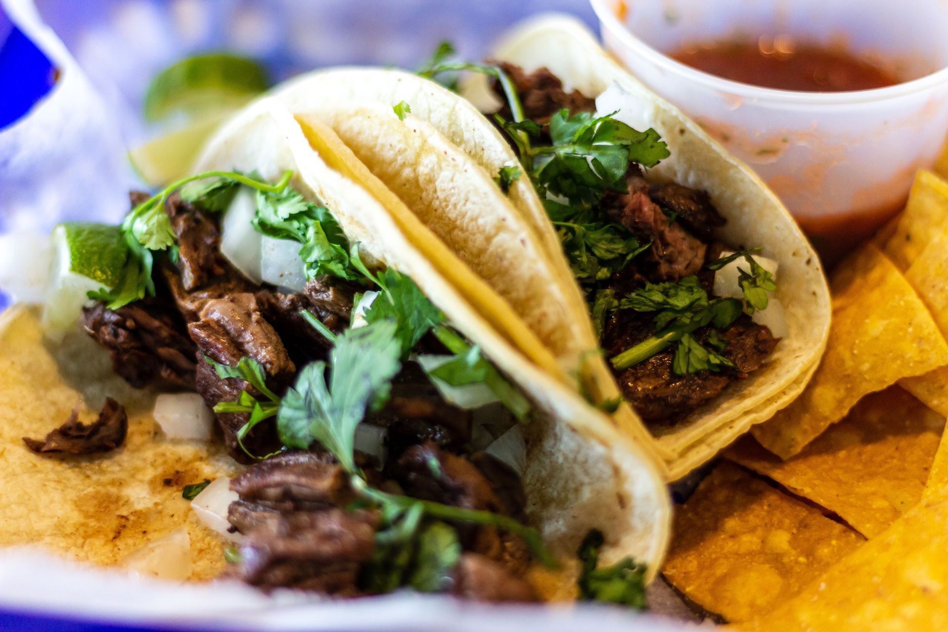 Two street-style steak tacos with cilantro and onion, served with tortilla chips and a side of salsa in a basket.