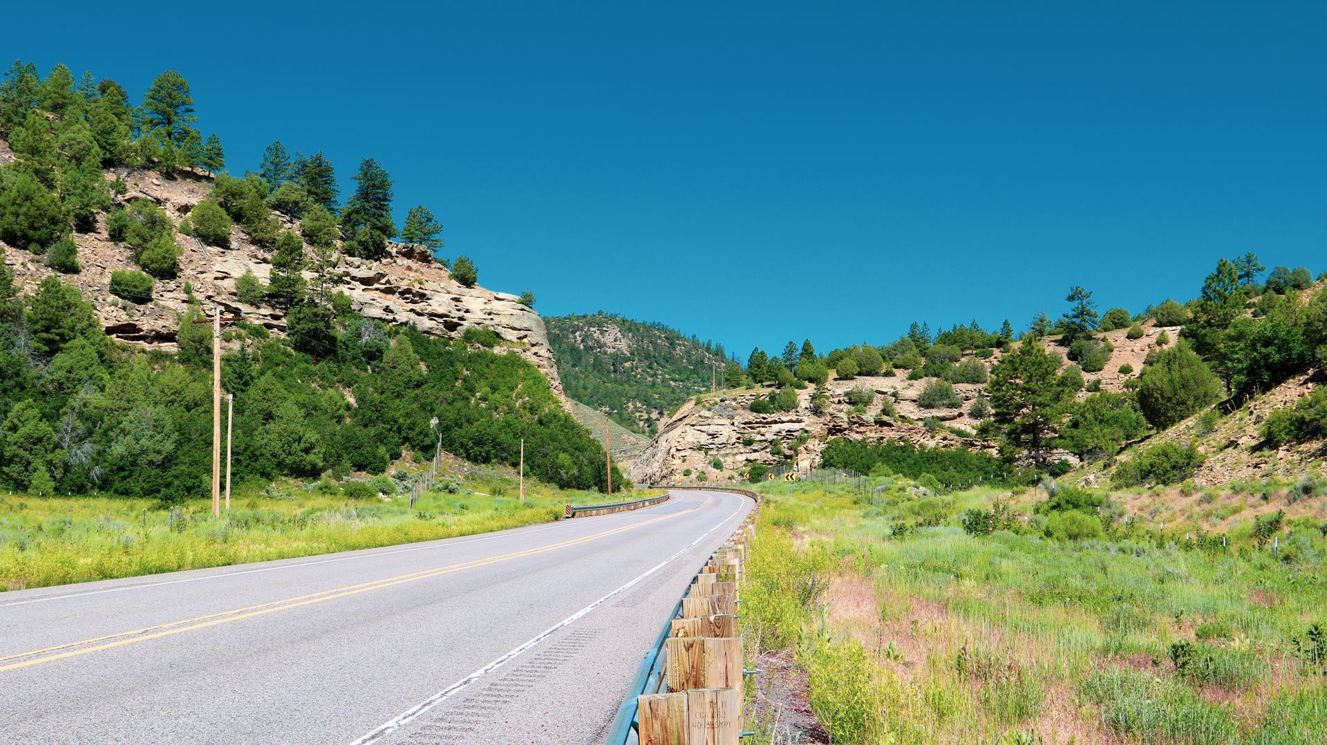 A paved road winds through a sunny canyon, flanked by rocky cliffs and green vegetation under a clear blue sky.