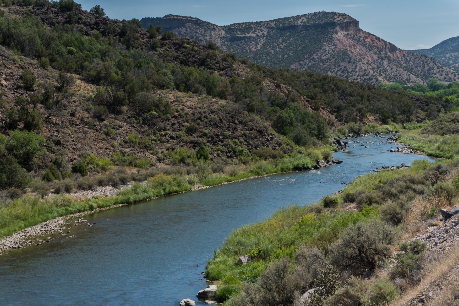 A river winds through a scenic canyon landscape with rocky cliffs, lush green vegetation, and shrubs under a blue sky.