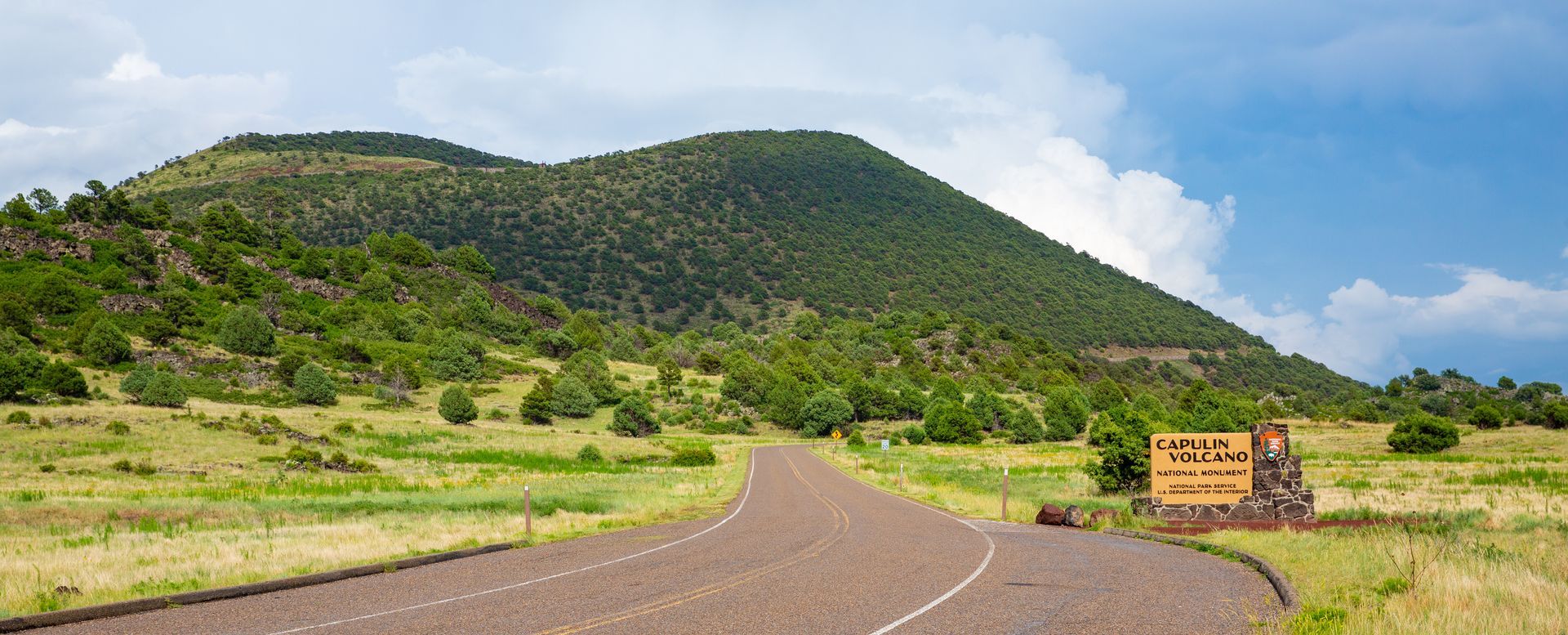 A paved road leads toward a green, hilly landscape under a blue sky with clouds, near a park entrance sign.