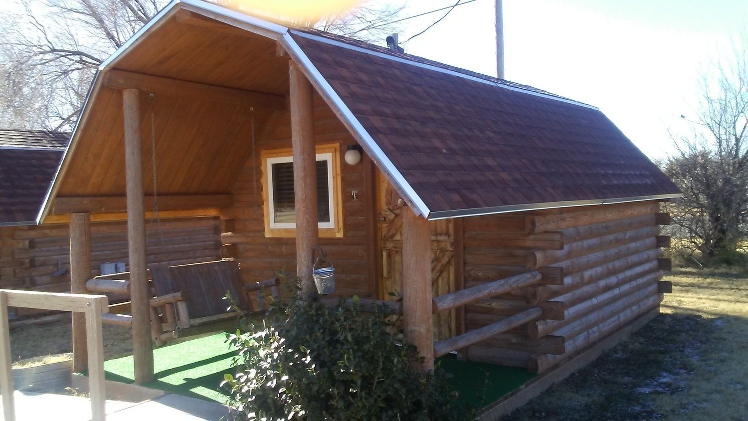 Small rustic wooden cabin with a covered porch, a swing seat, and a dark shingled roof, set in a sunny outdoor area.
