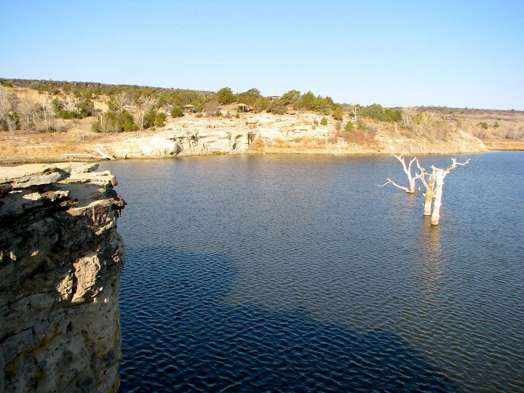 A rocky cliff overlooks a calm lake with two bare tree trunks emerging from the water under a clear blue sky.