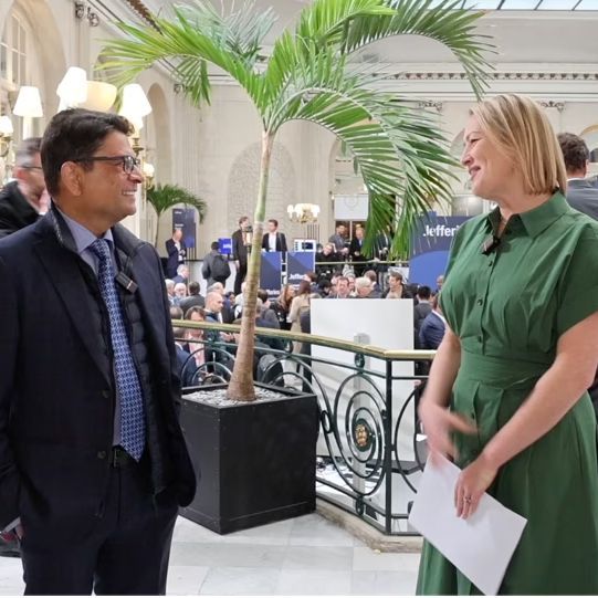 Man and woman conversing near a palm tree in a large hall with a crowd in the background.