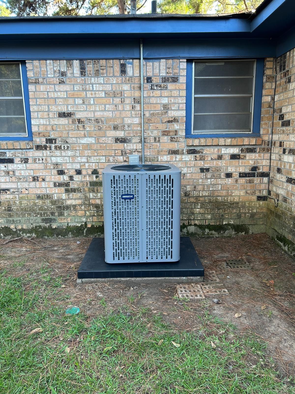 Air conditioner unit outside brick building. Gray unit on black base, with silver pipe extending upwards.