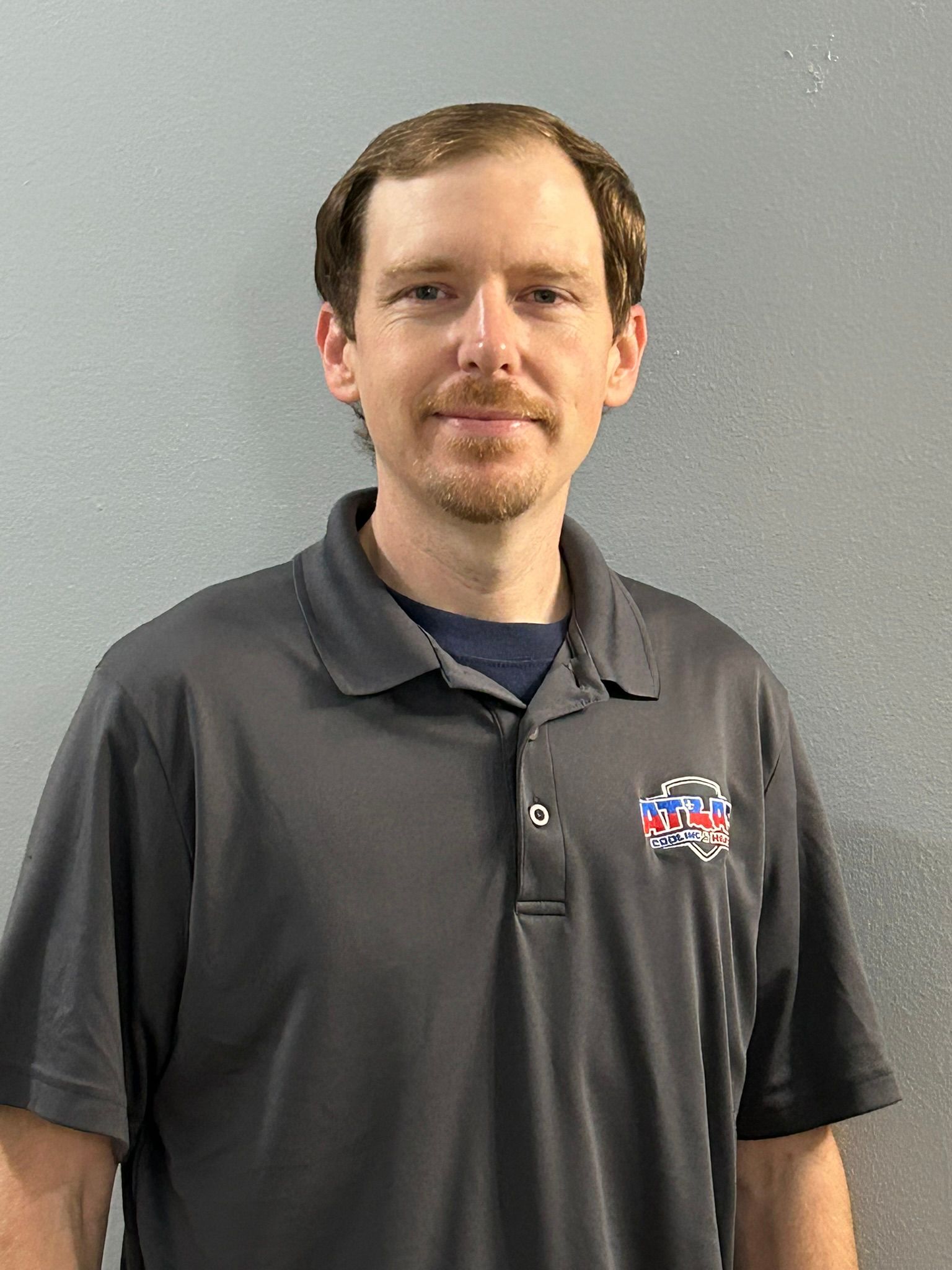 Man in gray polo shirt smiles in front of a gray wall.