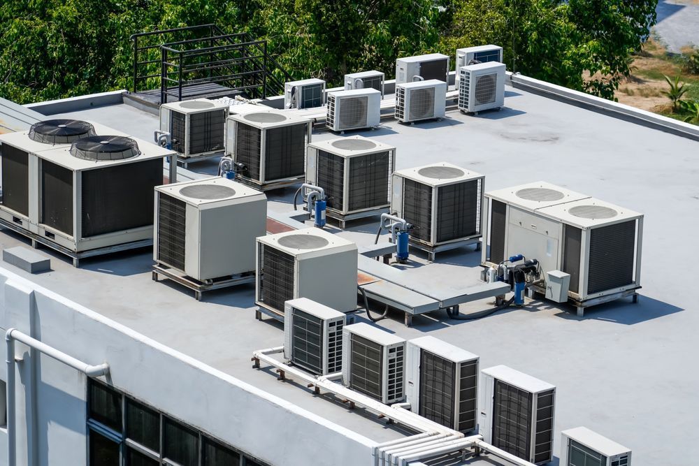 Rooftop with numerous air conditioning units, gray and black, against a backdrop of green trees.