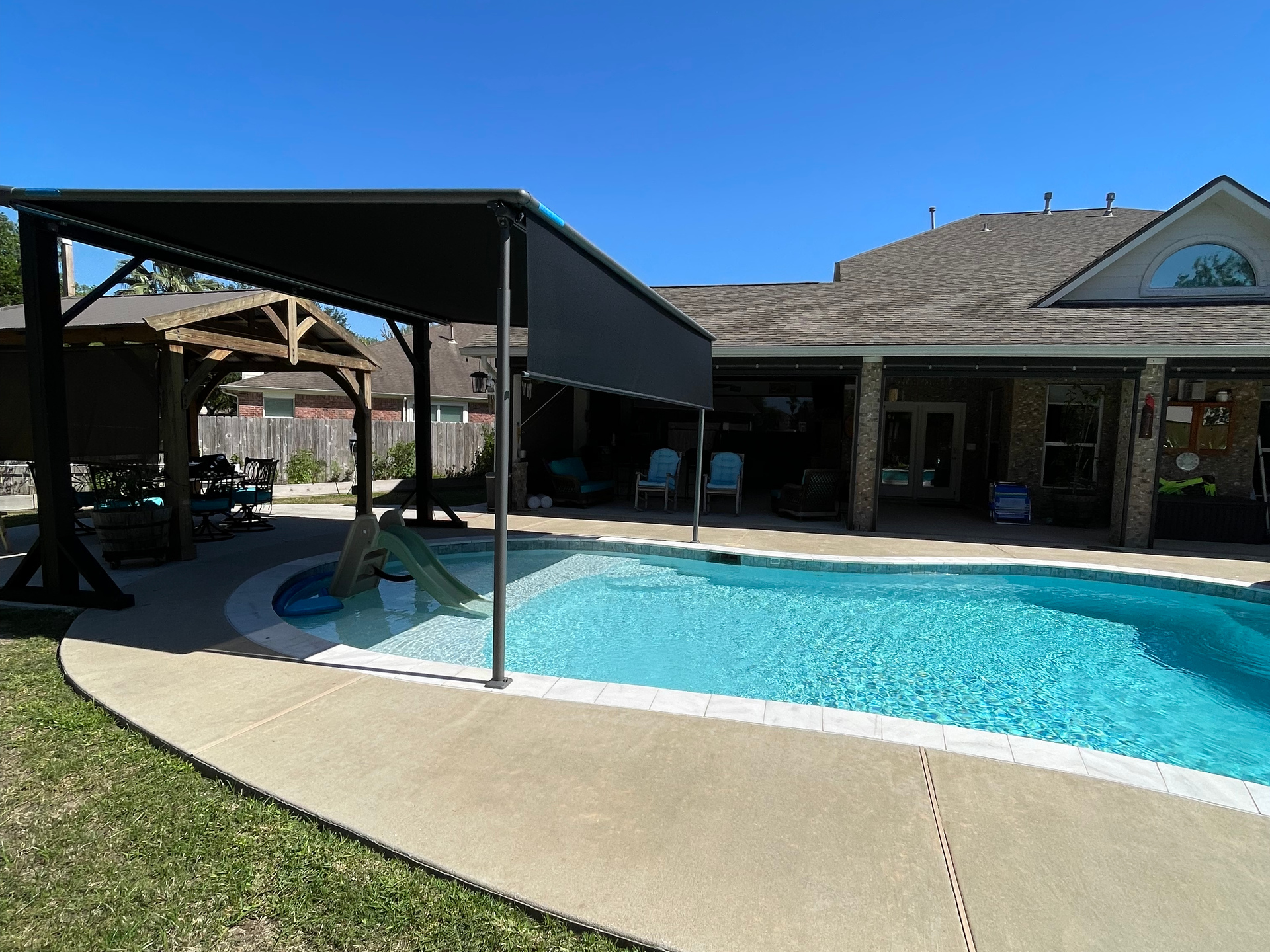 There is a large swimming pool in the backyard of a house with a motorized pergola