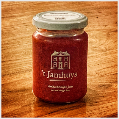 Een pot rode jam op een houten ondergrond, met een etiket waarop een huisillustratie en de tekst