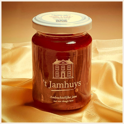 Een pot jam met een wit deksel en etiket, waarop een huis staat afgebeeld, rust op een beige stoffen ondergrond.