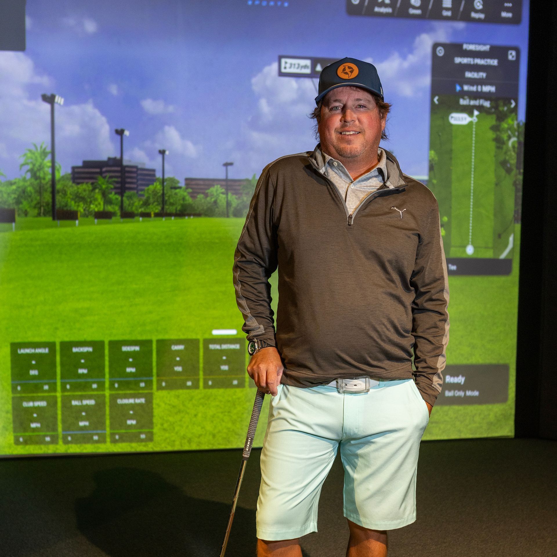 Man in shorts, holding golf club, in front of golf simulator screen.