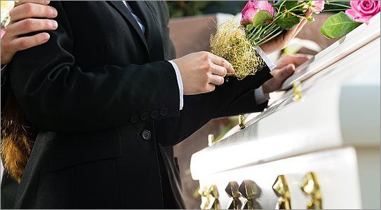 A woman standing with her hand on a white coffin, another arm comfortingly around her shoulder