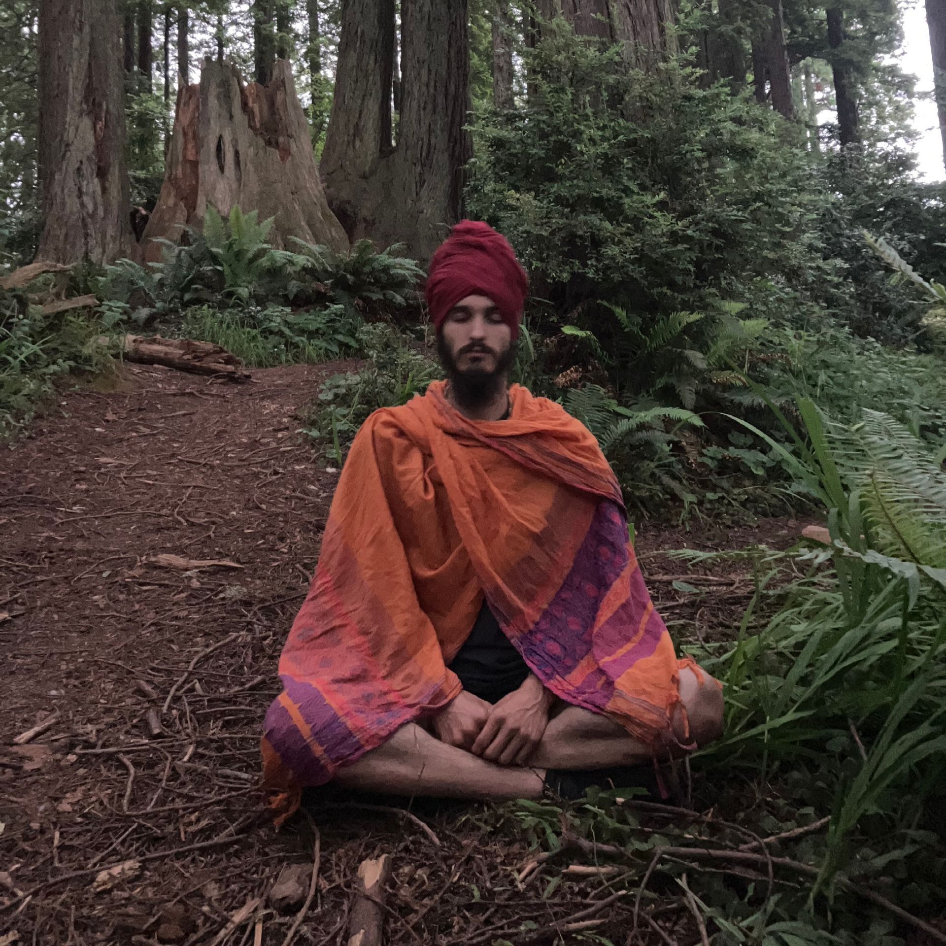 A man in a turban is sitting on the ground in the woods.