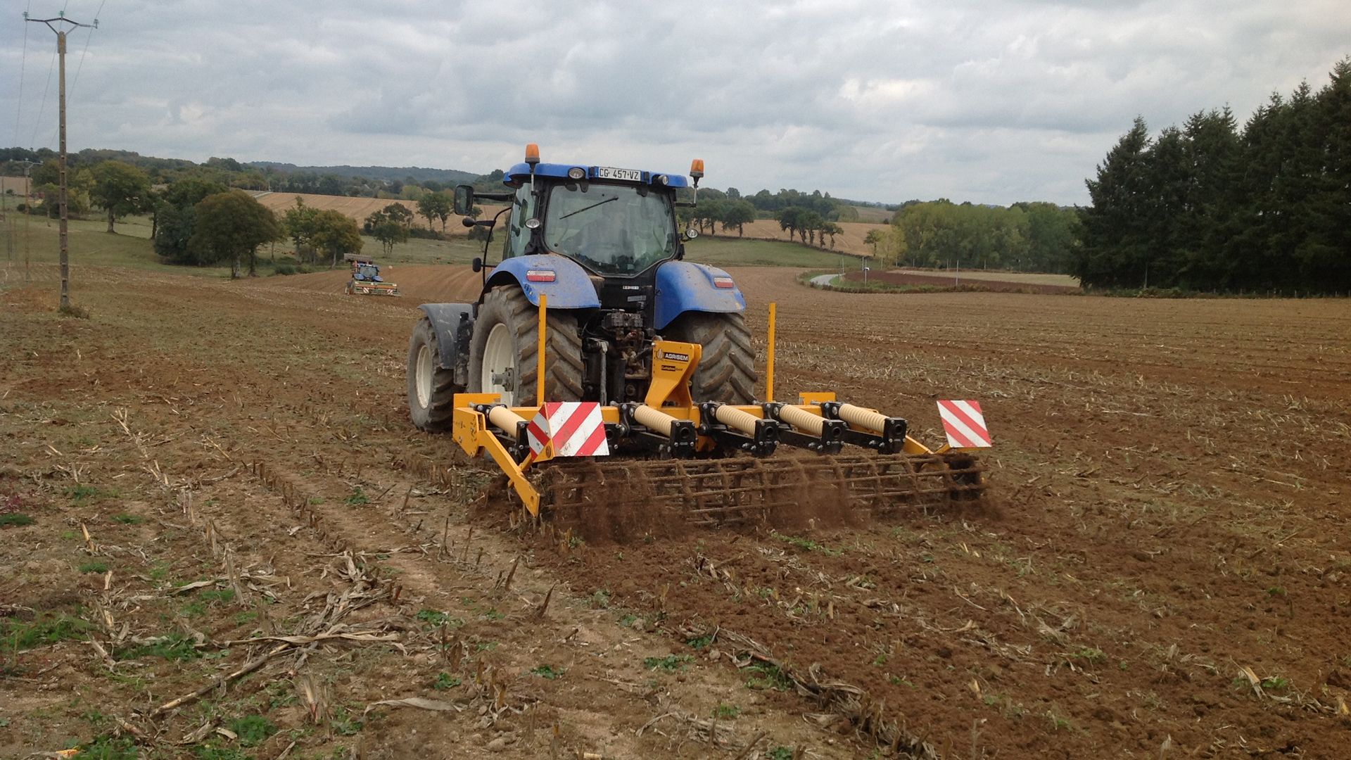 A blue and yellow tractor is towing a disc tiller in a field.
