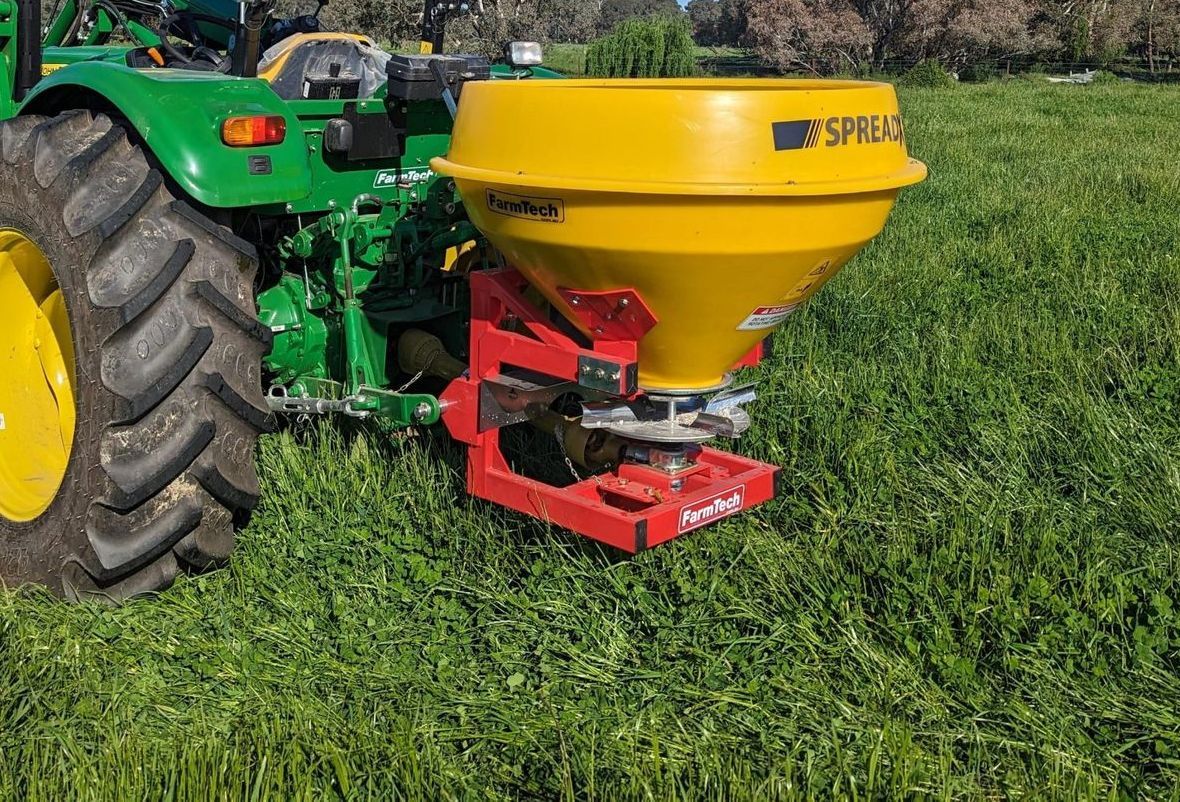 A green tractor with a yellow spreader attached to it is sitting in a grassy field.