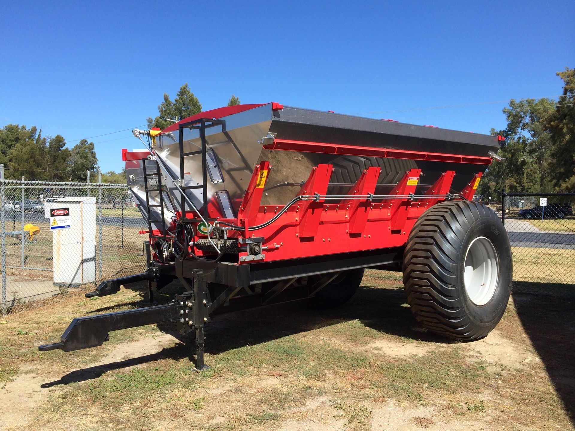 A red and black trailer is parked in the grass in front of a fence.