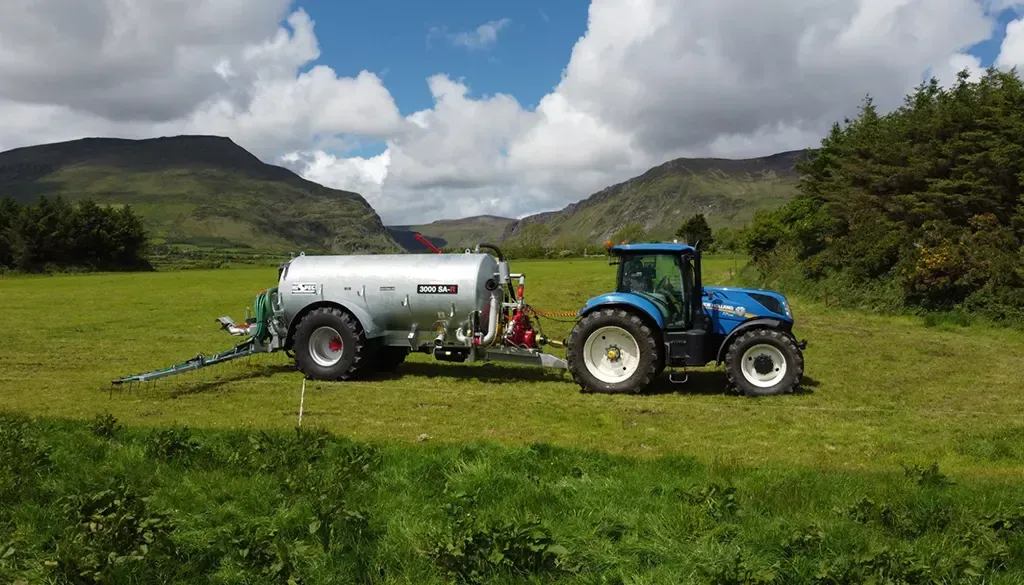 A blue tractor is driving through a grassy field with a trailer attached to it.