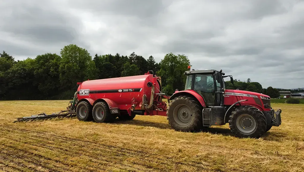 A red slurry tanker is driving through a field with a trailer attached to it.
