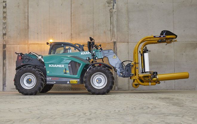 A green tractor with a yellow bale wrapper attached to it is parked in front of a concrete wall.